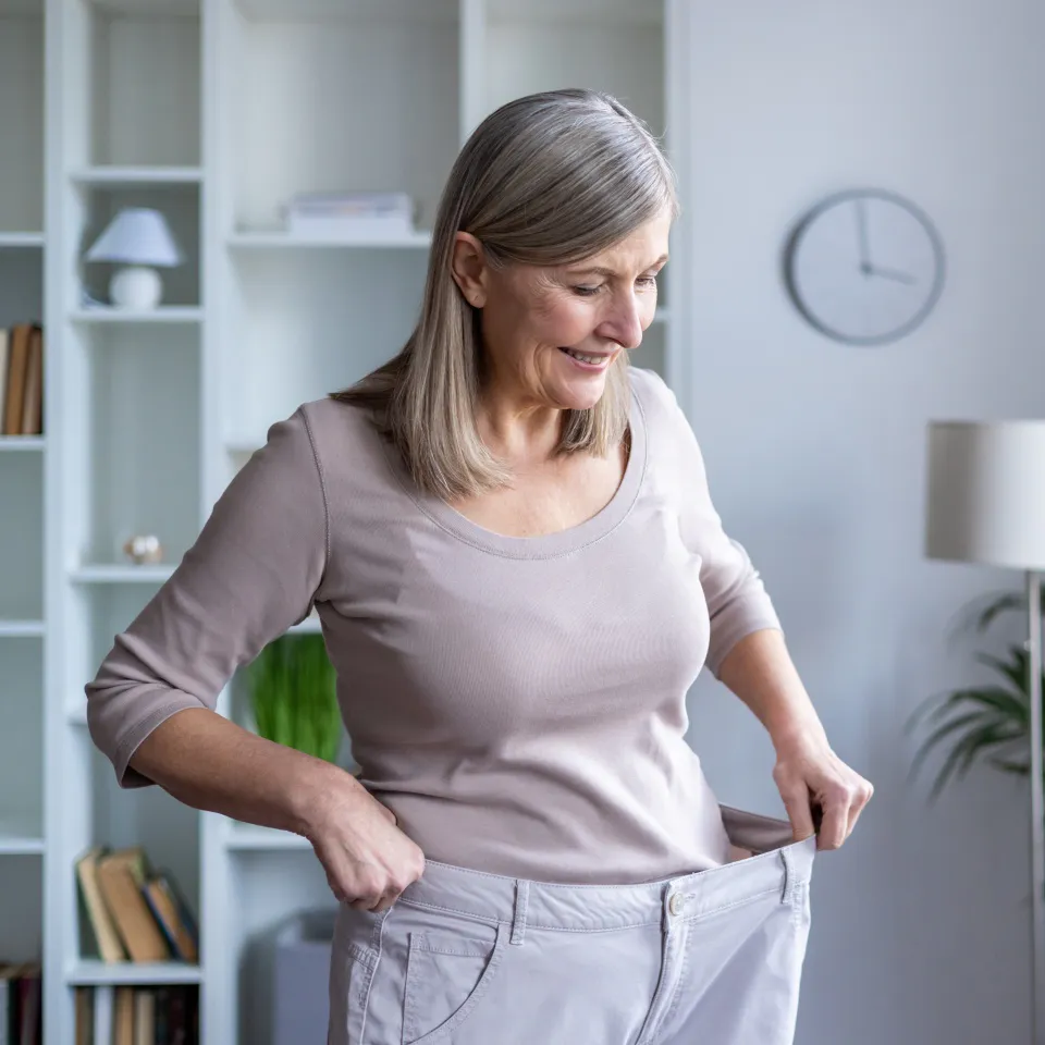 Older woman smiling and holding out the waistband of loose pants, suggesting weight loss, standing in a bright room with shelves, a plant, and a clock in the background.