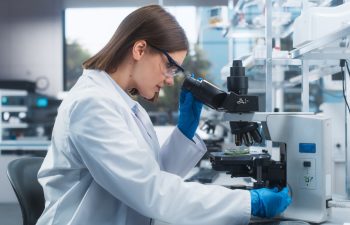 A person in a white lab coat and blue gloves examines a sample through a microscope in a laboratory setting.