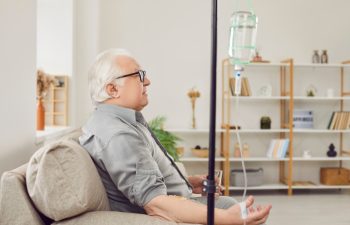 Senior man receiving vitamin therapy while resting on couch at home. Side view, profile portrait of old man sitting on sofa, drinking water and receiving medication solution through IV drip