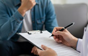 A person in a denim jacket sits on a couch while a healthcare professional writes on a clipboard during a consultation.