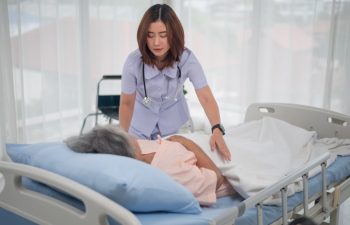 Asian nurse in nursing home support old man for sleeping on the bed in patient room, Compassionate nurse assistant offering gentle care to senior woman lying in bed.