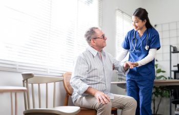 A friendly female nurse in blue scrubs stands beside an elderly male patient, offering him comfort and support. The clinic or home setting suggests a moment of compassionate healthcare,