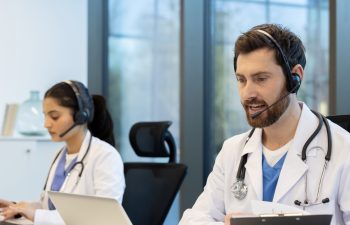 Medical professionals wearing headsets and lab coats, sitting at desks with laptops, are providing remote online healthcare consultation services in a modern office helping patients