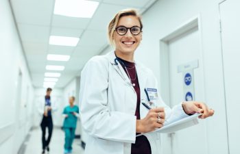 Portrait of happy young female physician standing in corridor with clipboard. Caucasian woman working in hospital writing prescription.