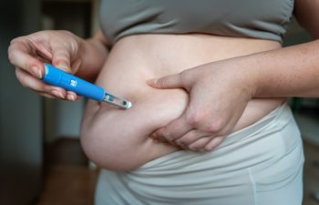 woman injecting a GLP-1 weight loss medication into belly fat, demonstrating semaglutide treatment at home. The image shows a hand holding a blue injection pen and pinching abdominal skin, illustrating obesity therapy, fat reduction and modern metabolic healthcare.