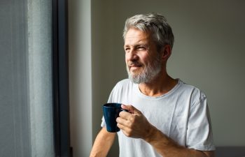 A middle-aged man with gray hair and a beard stands by a window holding a blue mug, looking outside. He is wearing a plain white t-shirt.