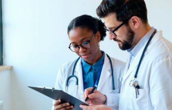 Black young adult woman and Caucasian young adult man doctors reviewing medical chart together, both wearing stethoscopes and glasses, collaborating in clinical setting