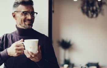 Adult man in casual wear resting in cafe. He standing in with mug of tea against blurred background and smiling