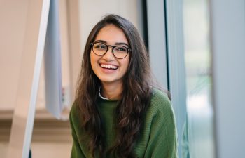 A woman with long dark hair and glasses, wearing a green sweater, smiles while sitting indoors near a window.