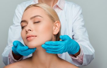 cropped view of beautician in blue latex gloves touching neck of naked woman with closed eyes isolated on grey