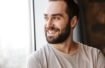 Handsome smiling young bearded man standing at the window at home, looking away