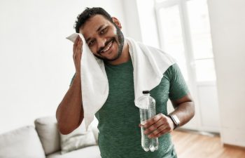 Tired but happy. Young man smiling at camera, holding water bottle and wiping sweat with towel while resting after training workout at home. Sport, healthy lifestyle. Horizontal shot