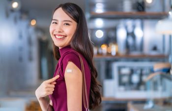 A woman smiles at the camera while showing a small adhesive bandage on her upper arm, indicating she has just received a vaccination.