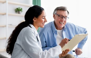 Young doctor asking senior impaired male patient in wheelchair to sign insurance policy at home. Handicapped elderly man putting his signature under surgery consent form, reading medical document