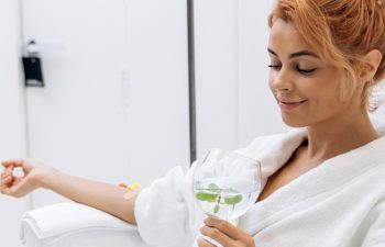Waist up portrait view of the charming woman in white bathrobe sitting in armchair and receiving IV infusion. She is holding glass of lemon beverage and smiling