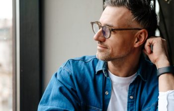 Adult handsome man in glasses looking out window and touching his head while sitting in cafe with phone