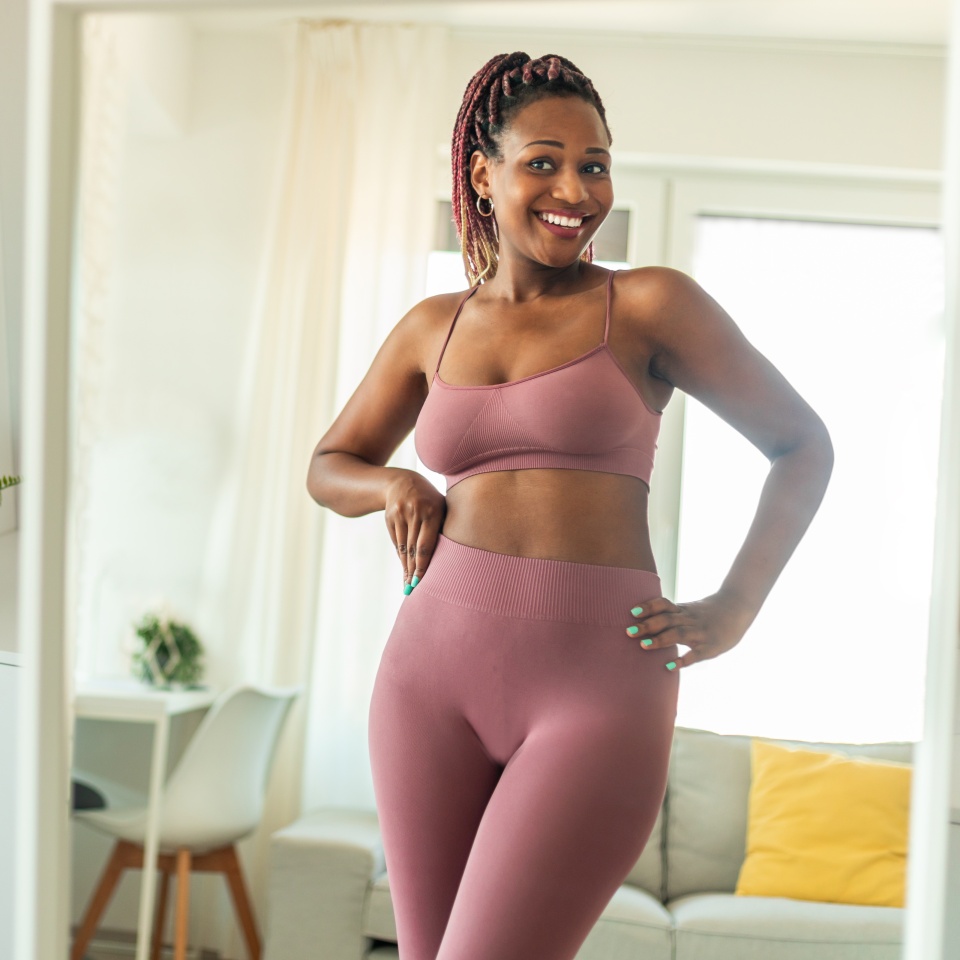 Successful weight loss concept. Happy slim african american woman in sports clothes looking at her reflection in mirror and smiling, feeling satisfied with slimming
