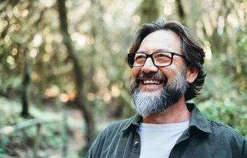 Portrait of young mature man alone smiling outside with nature trees in background. Environment. Happy people enjoying outdoor leisure activity. Nature feeling. Bearded adult male wearing eyeglasses