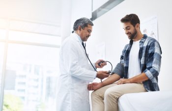 Healthcare, patient and doctor with blood pressure test, help and wellness checkup of person at hospital. Medicine, health care expert and Indian man in doctors office consulting medical professional.