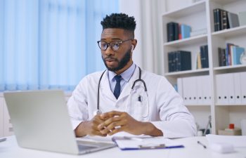 A doctor wearing a white coat and stethoscope sits at a desk, looking at a laptop, with medical files and books in the background.