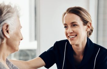 Senior woman, nurse and smile with support for healthcare, empathy or trust in elderly care or retirement at home. People, caregiver and touching person with a disability for help, love and comfort.