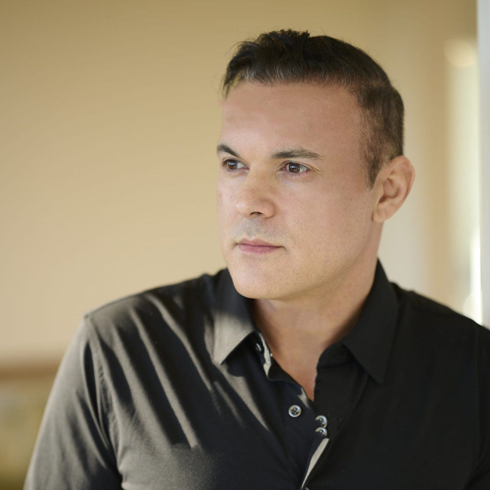 A man with short dark hair wearing a black button-up shirt looks to the side in a softly lit indoor setting.