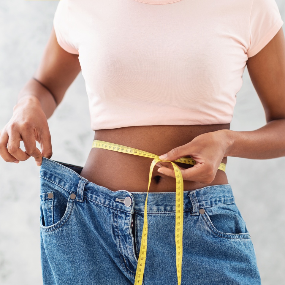 Closeup of black woman in old big jeans measuring her waist with tape, showing results of slimming diet or liposuction, grey background. Young lady promoting healthy nutrition for weight loss