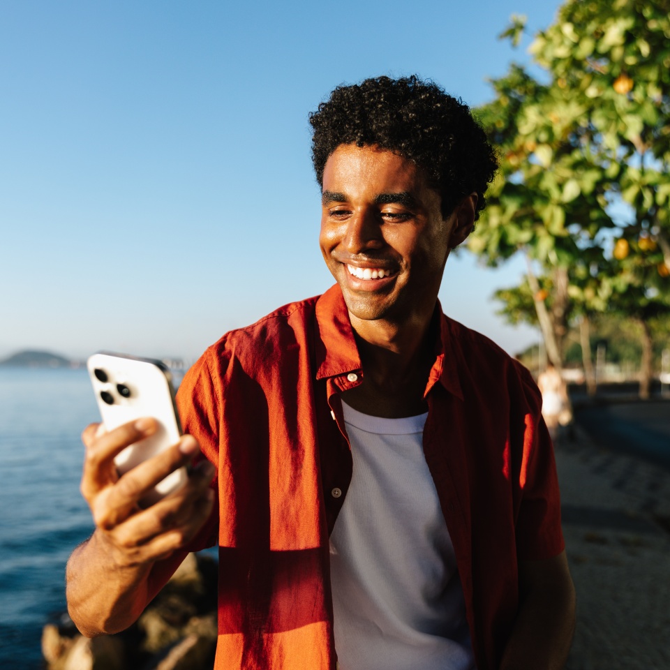 A young man smiles while using his smartphone along Urca's Wall, with a scenic view of the ocean. The clear blue sky adds to the relaxed mood, capturing a perfect blend of technology and nature.