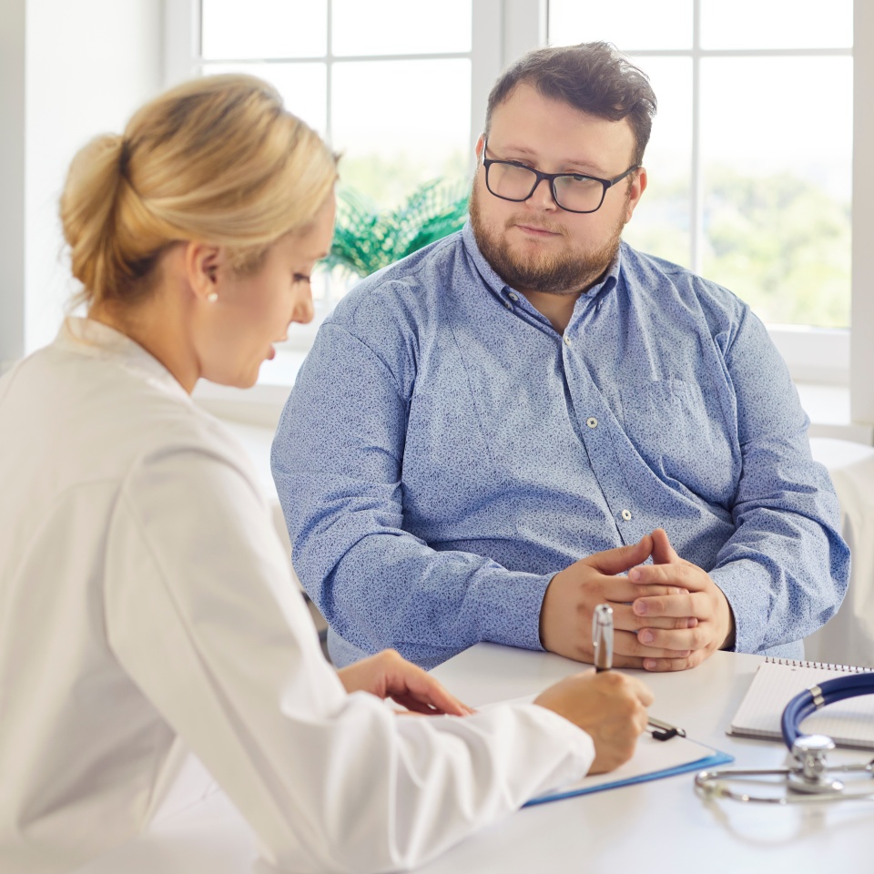 Doctor and patient during obesity consultation support. The physician writes notes as the man listens, stethoscope on the desk by the window. Collaborative medical weight management and counseling.
