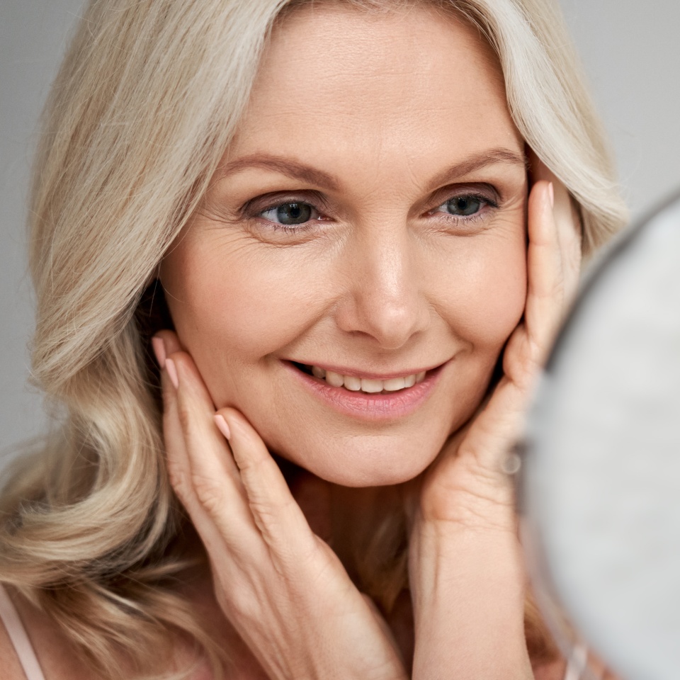 Happy 50s middle aged woman model touching face skin looking in mirror. Smiling mature older lady pampering, enjoying healthy skin care, aging beauty, skincare treatment cosmetic products concept.