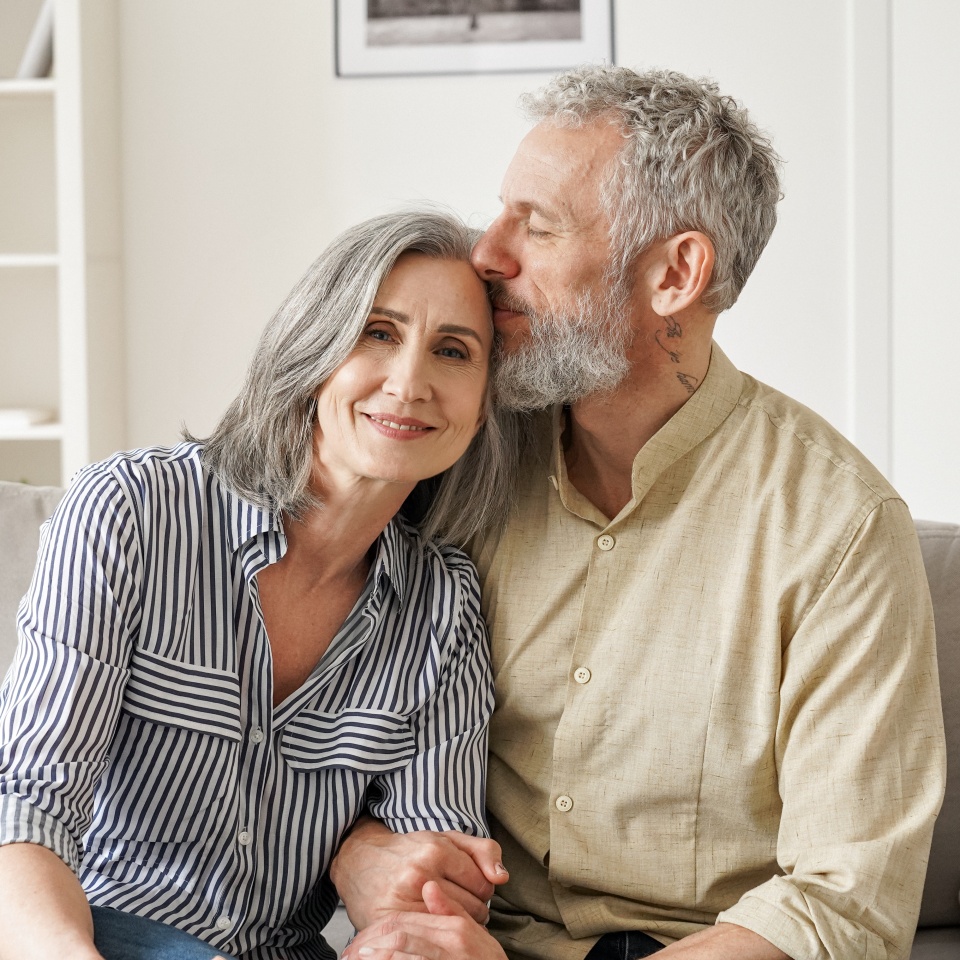 Happy affectionate classy older mature couple bonding with eyes closed at home. Loving caring senior 50s husband embracing and kissing mid aged wife enjoying tender moment sitting on couch at home.