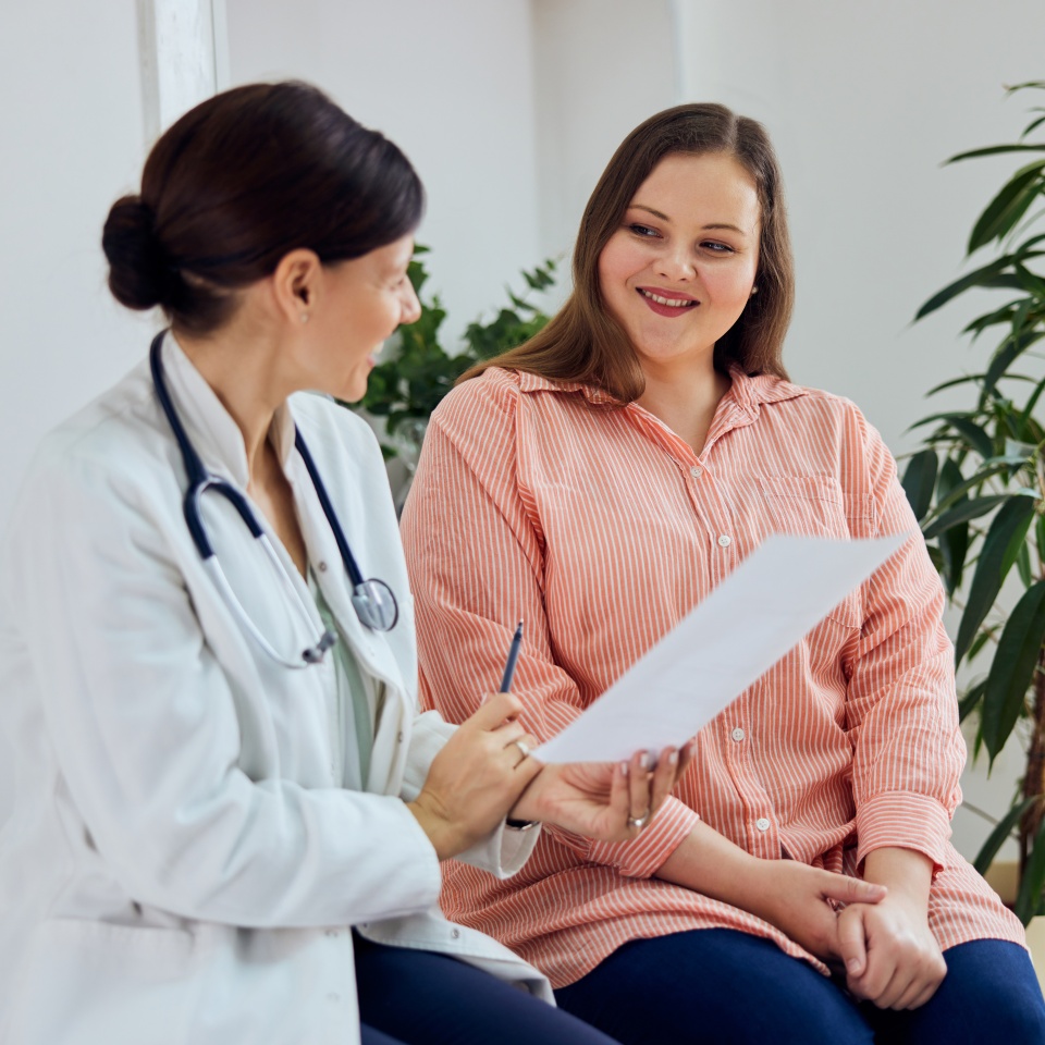 A satisfied female nutritionist holding a paper with weight loss progress and smiling with an overweight female patient.